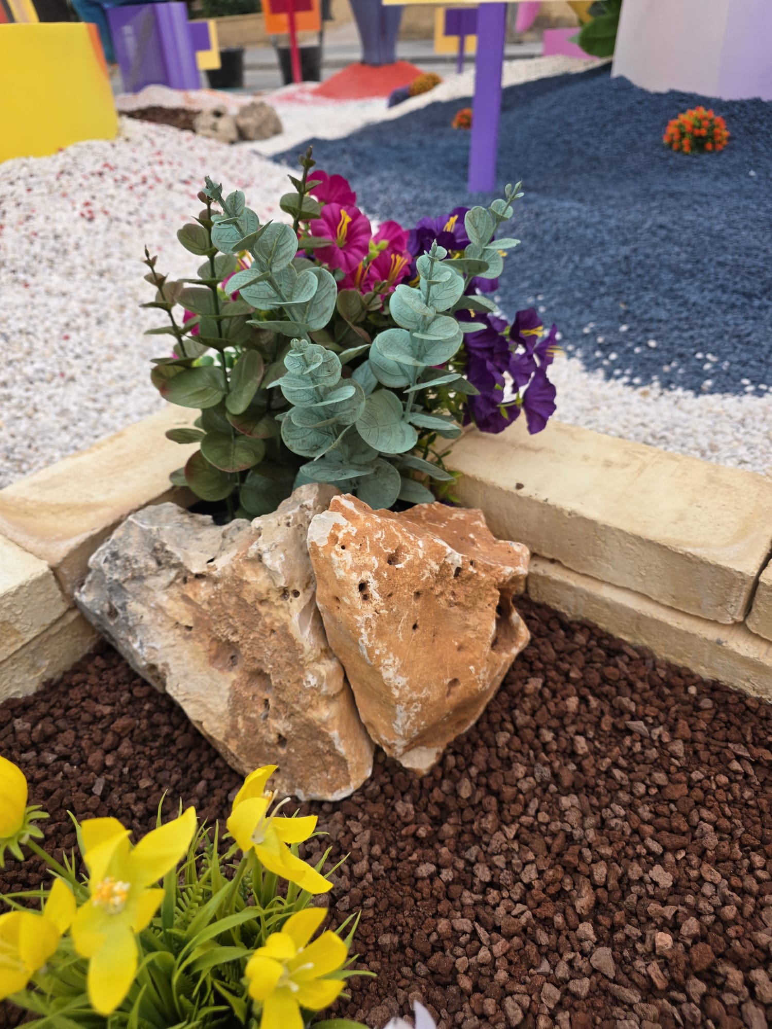 Detalle de la decoración de la Hoguera Grande Princesa Mercedes 2026, donde la rocalla natural se combina con plantas artificiales y piedra volcánica roja para crear una composición equilibrada, aportando textura, volumen y un acabado paisajístico integrado en la ambientación.